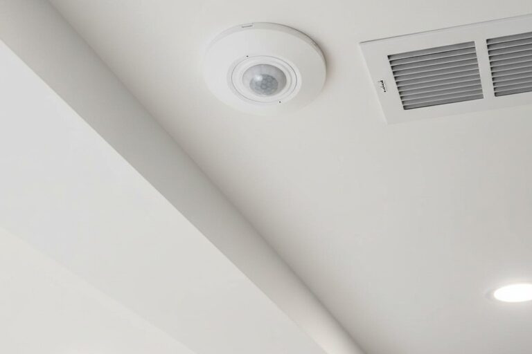 Uma foto de ângulo baixo olhando para cima, mostrando um pequeno sensor de movimento branco e redondo embutido no teto de drywall, posicionado ao lado de uma grelha de ar de metal quadrada.