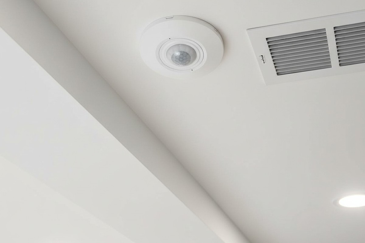 Uma foto de ângulo baixo olhando para cima, mostrando um pequeno sensor de movimento branco e redondo embutido no teto de drywall, posicionado ao lado de uma grelha de ar de metal quadrada.