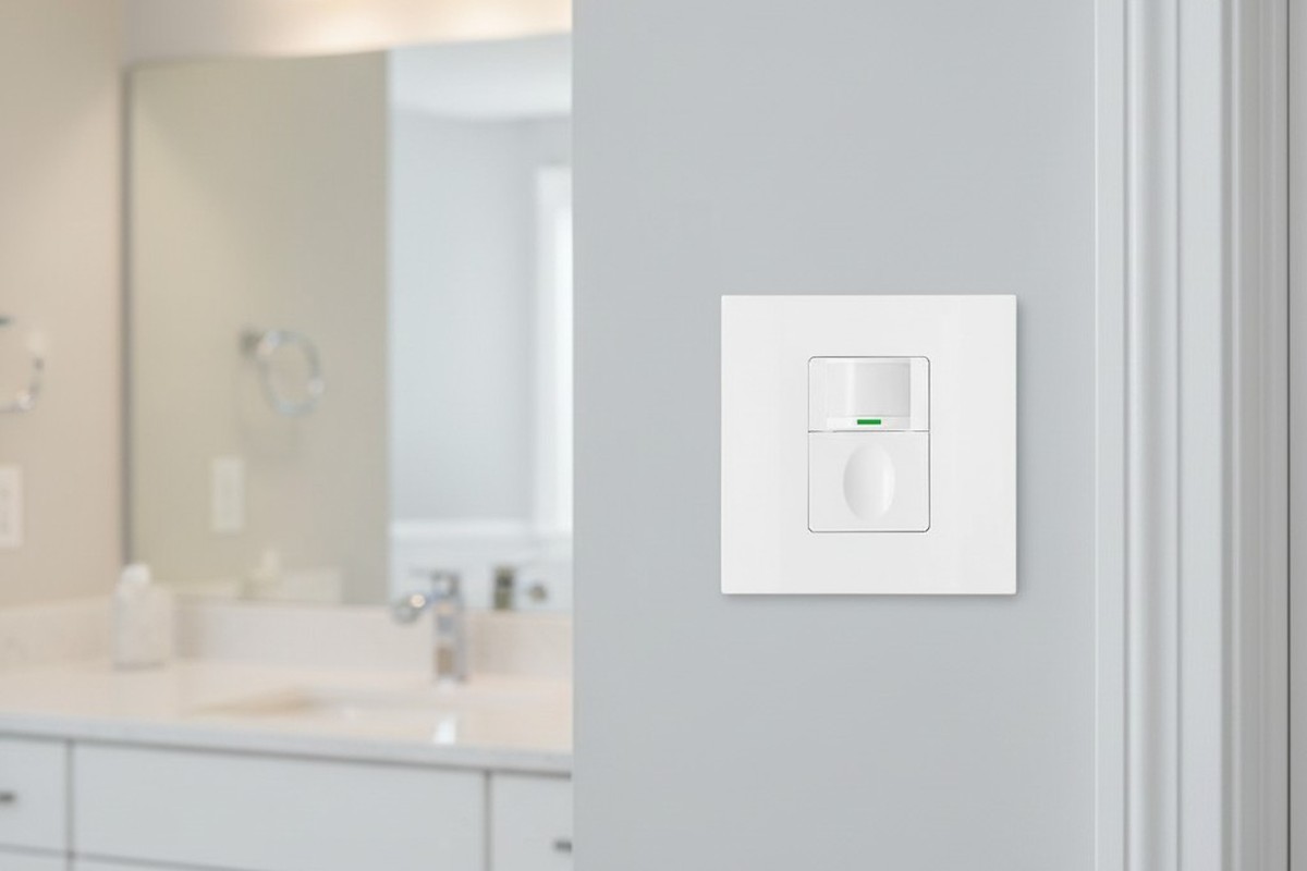Um interruptor de sensor de vaga em branco está instalado de forma organizada em uma parede cinza claro ao lado de uma moldura de porta em um banheiro moderno e limpo, com uma bancada desfocada ao fundo.