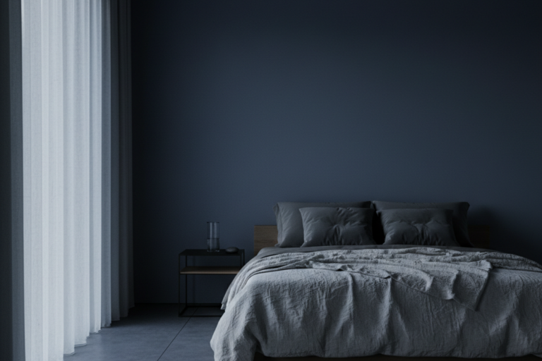 Um quarto pouco iluminado apresenta uma cama baixa com roupas de cama cinza e um copo de água em uma mesa lateral contra paredes azul-escuras. Luz suave filtra através de cortinas transparentes do chão ao teto, iluminando o ambiente sem luminárias artificiais.