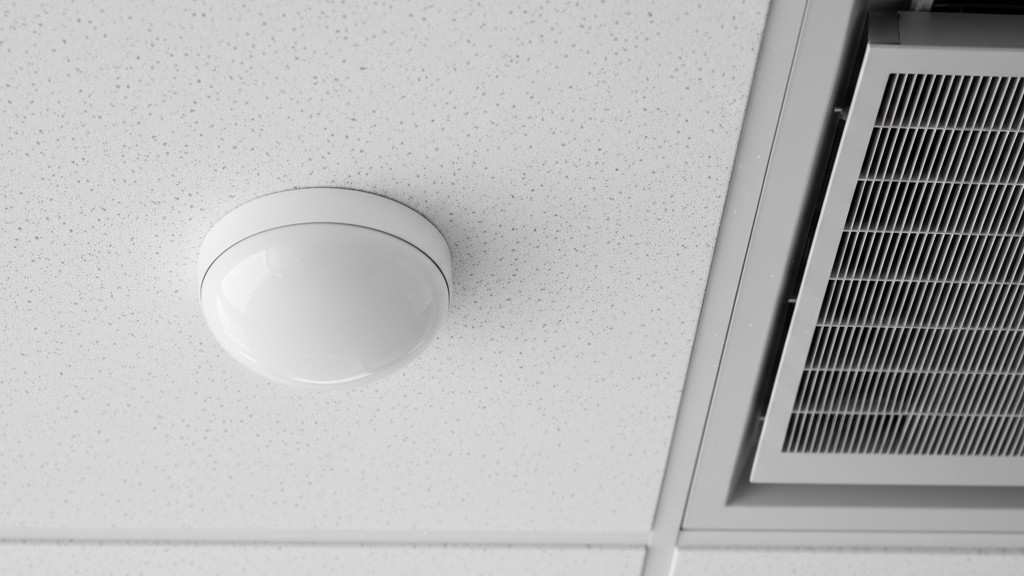 Um close-up de um sensor de movimento redondo branco no teto montado diretamente ao lado de uma saída de ar quadrada de metal do HVAC.