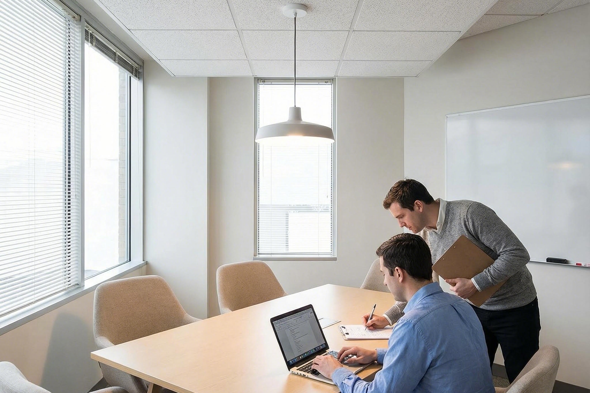 Test de présence assise dans une salle de réunion informelle Une petite table de salle de conférence avec des ordinateurs portables et une personne assise pendant qu'une autre marque une carte simple sur un clipboard sous un capteur au plafond.